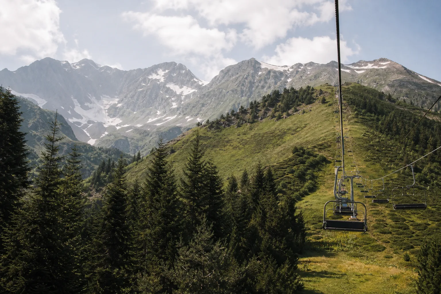 An open chairlift rising over spruce forest towards the grassy Zuruldi ridge above Mestia, with snow peaks behind