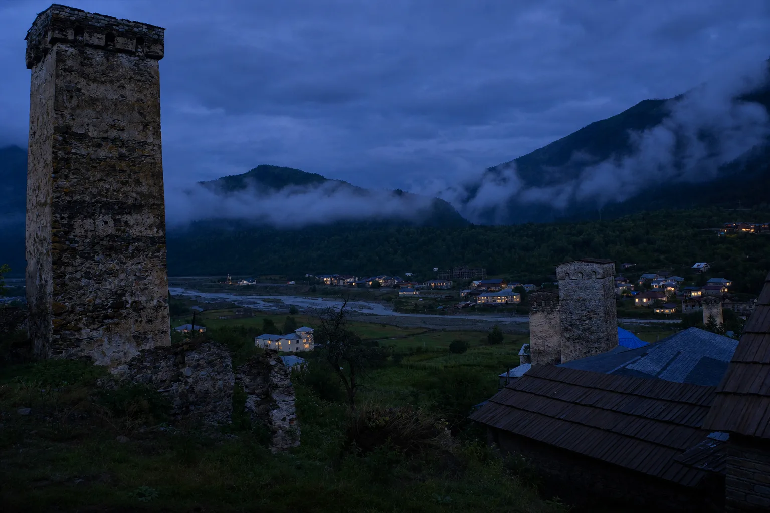 Stone Svan towers and slate-roofed houses of Mestia at dusk with a few lit windows and the dark ridge behind