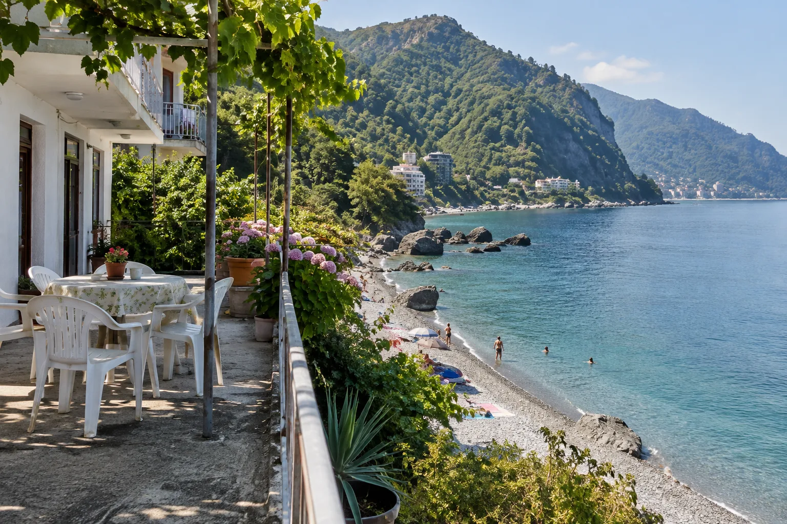 Family guesthouse terrace with plastic chairs above a pebble beach and clear sea at Kvariati, steep green hills behind