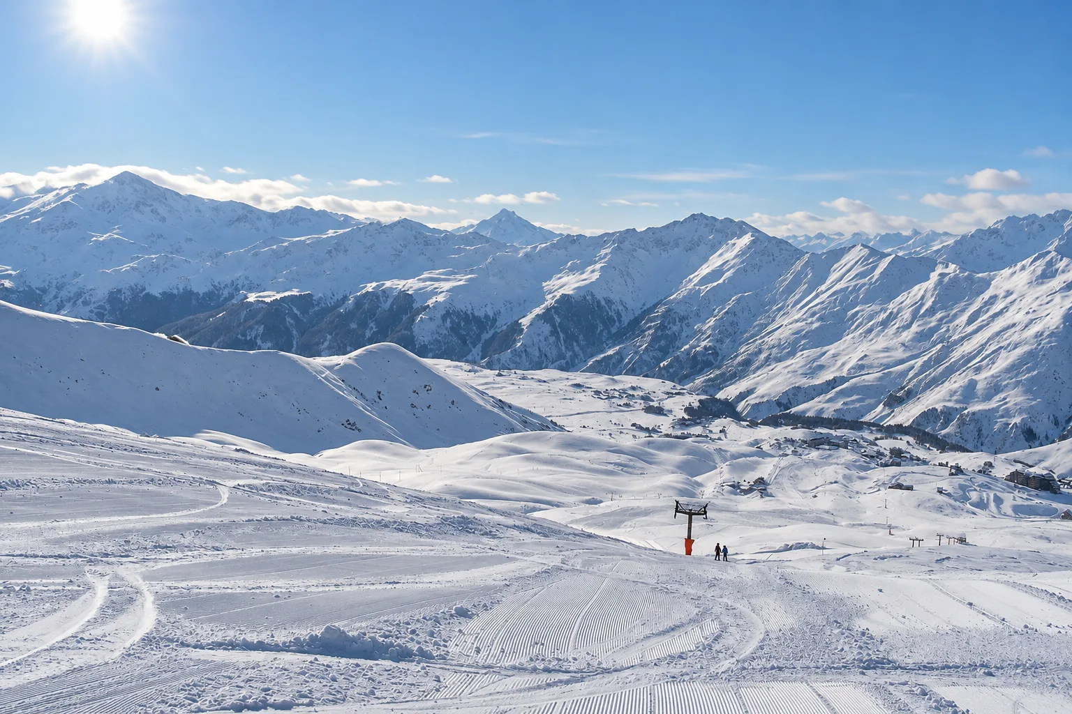Groomed ski slopes above Gudauri on a bright January afternoon with a chairlift pylon, two distant skiers and the resort buildings far below in a wide treeless valley of the Greater Caucasus
