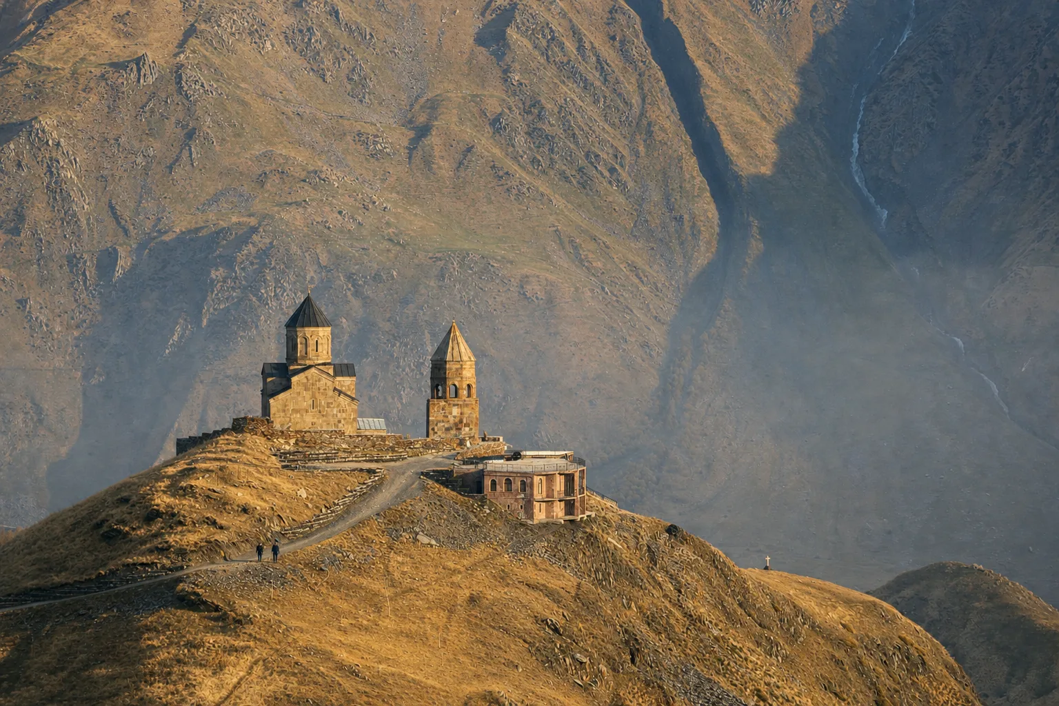 Gergeti Trinity Church and its bell tower on a grassy ridge above Stepantsminda in October, with the mountainside rising behind