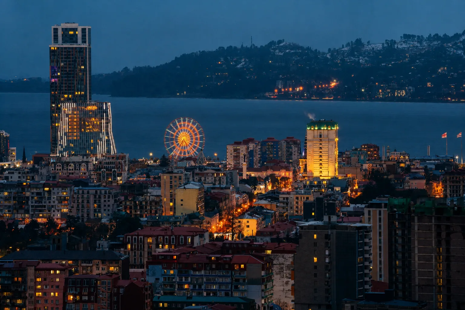 Batumi's towers and ferris wheel lit at dusk, seen from above, with the sea and the hills of the coast behind