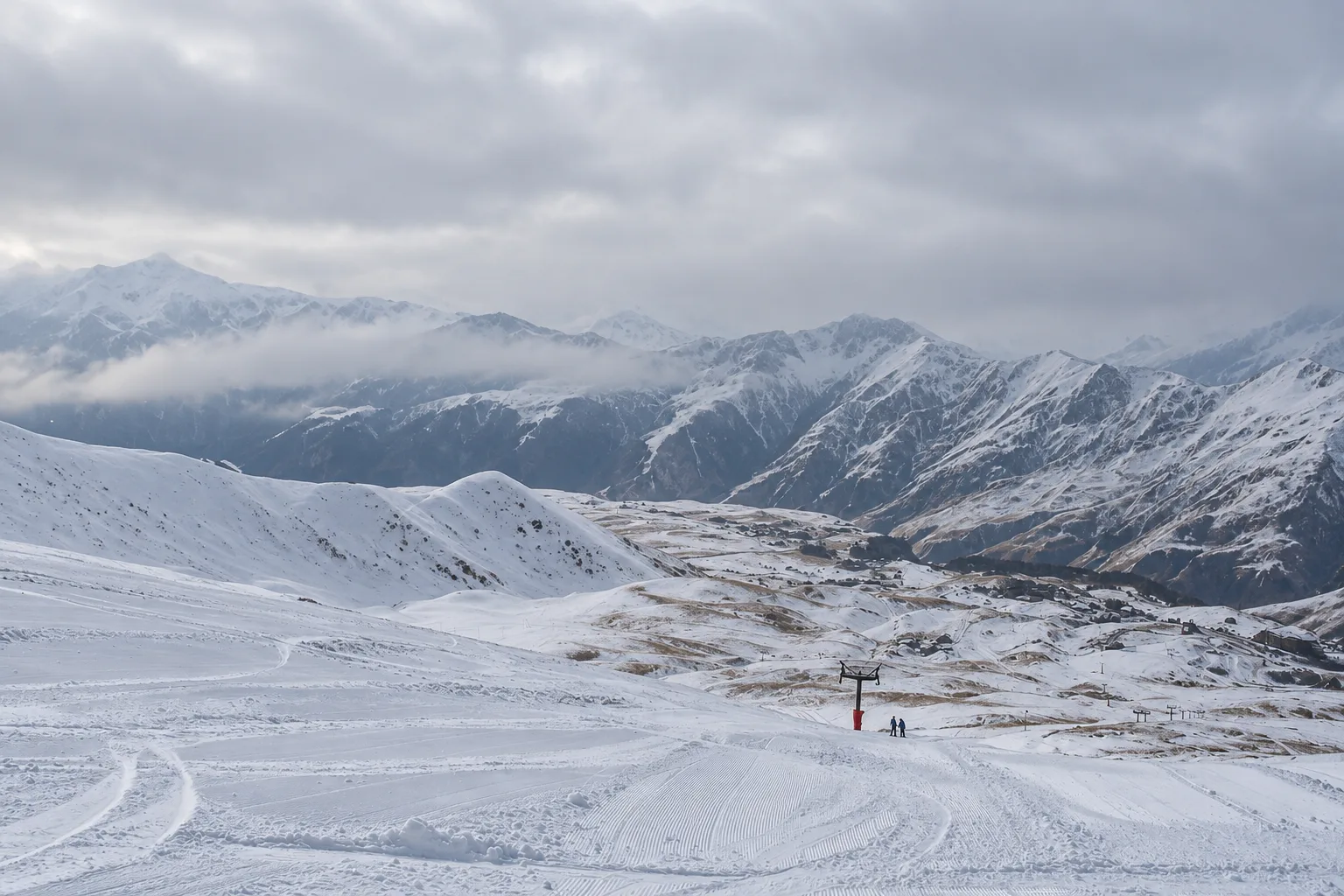 Groomed piste at Gudauri in late December with fresh snow on the upper slopes, thin cover and grass showing lower down, a lift pylon and two distant skiers