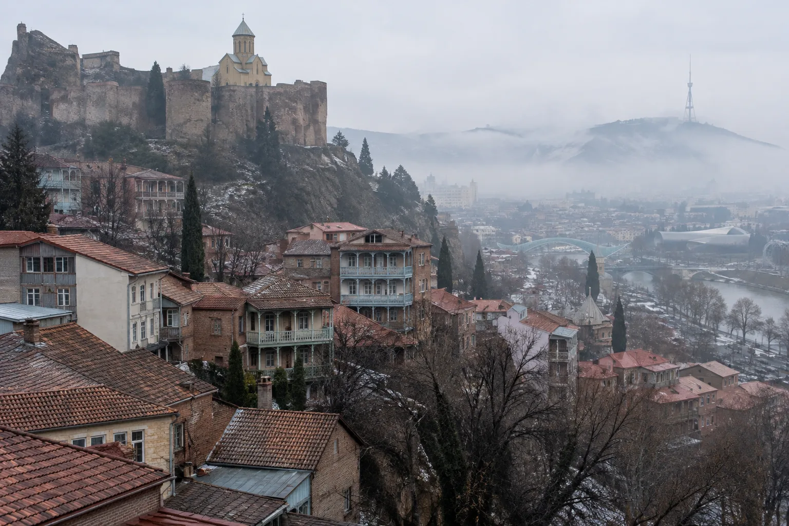 Tiled roofs and wooden balconies of Tbilisi's old town on a foggy January morning with the walls of Narikala fortress on the ridge above and the Mtkvari valley lost in fog
