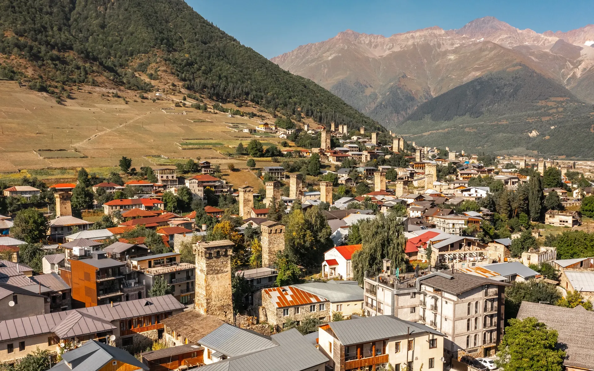 Aerial view of Mestia town in Svaneti with medieval stone towers and snow-capped Caucasus mountains under a blue sky