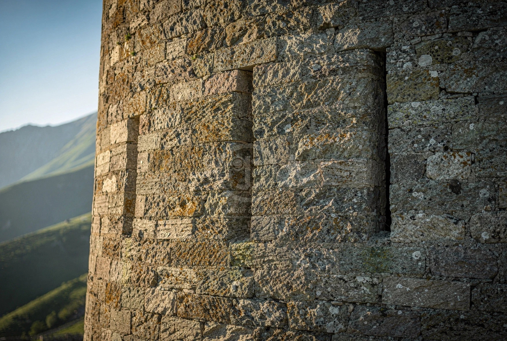 Close-up of ancient stone defensive tower walls with narrow arrow slits, rough-hewn masonry and mountain peaks visible behind