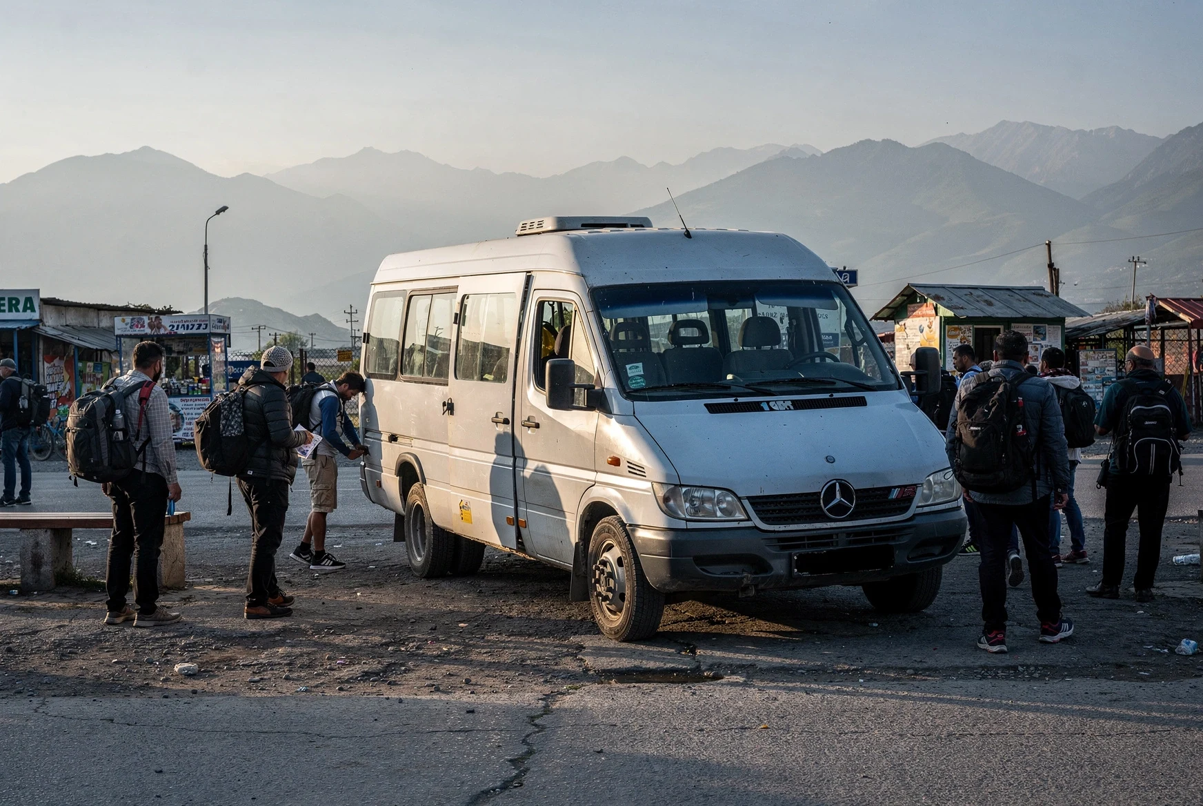 White intercity marshrutka minibus parked at a busy roadside transport stop in Georgia with mountains in the background