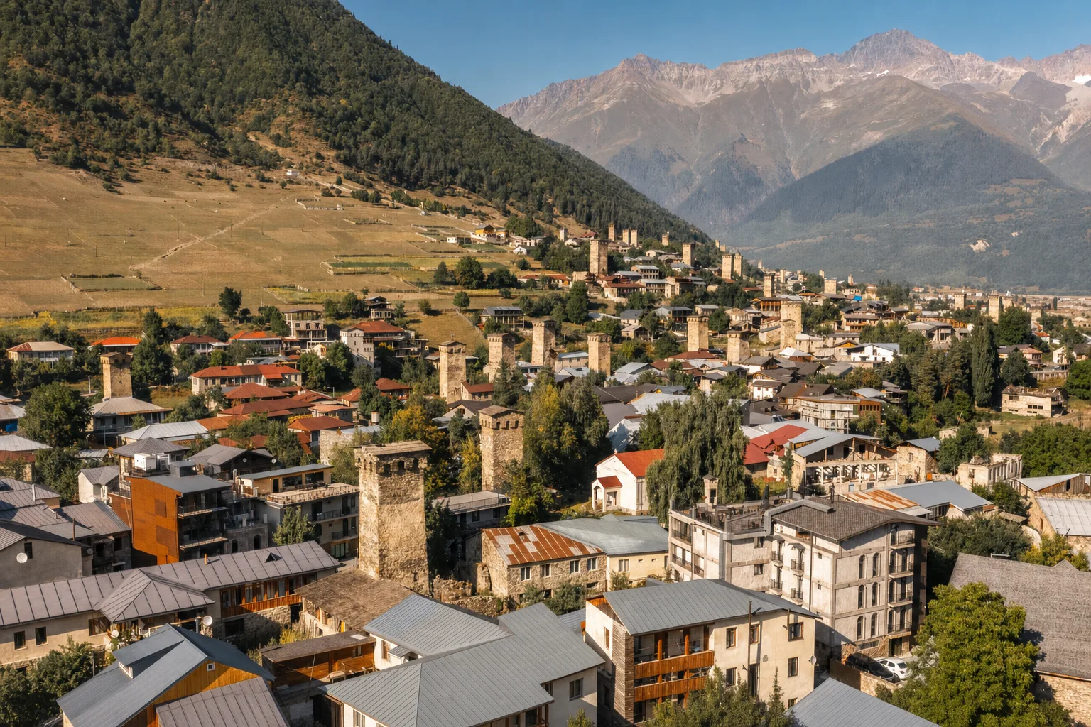 Mestia from above on a clear morning, stone towers and slate roofs filling the valley floor with forested slopes and snow peaks behind
