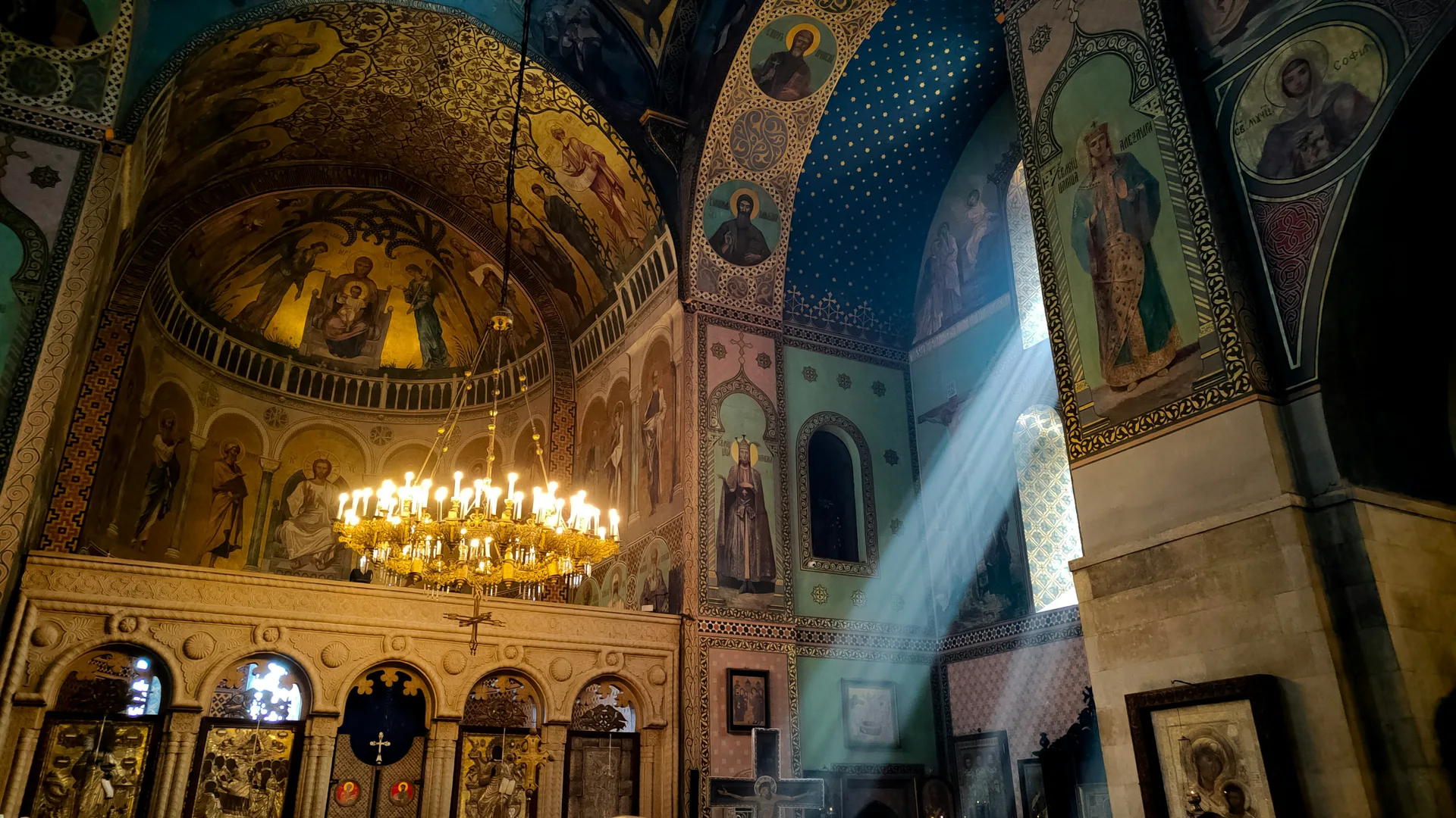 Interior of a Georgian Orthodox cathedral with golden frescoes, blue starry vaults, and shafts of light