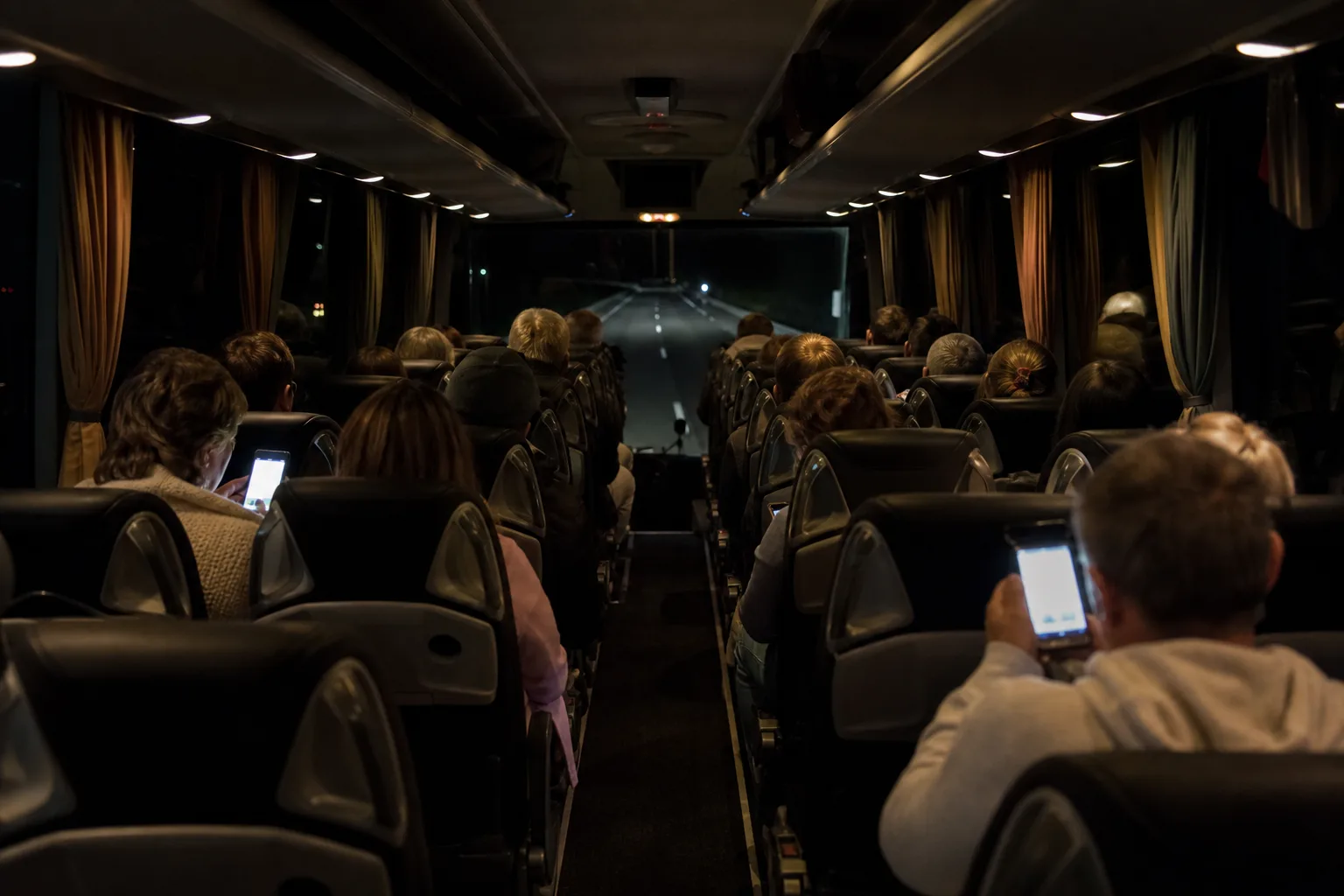Passengers seen from behind inside a modern intercity coach at night with a dark motorway through the windscreen