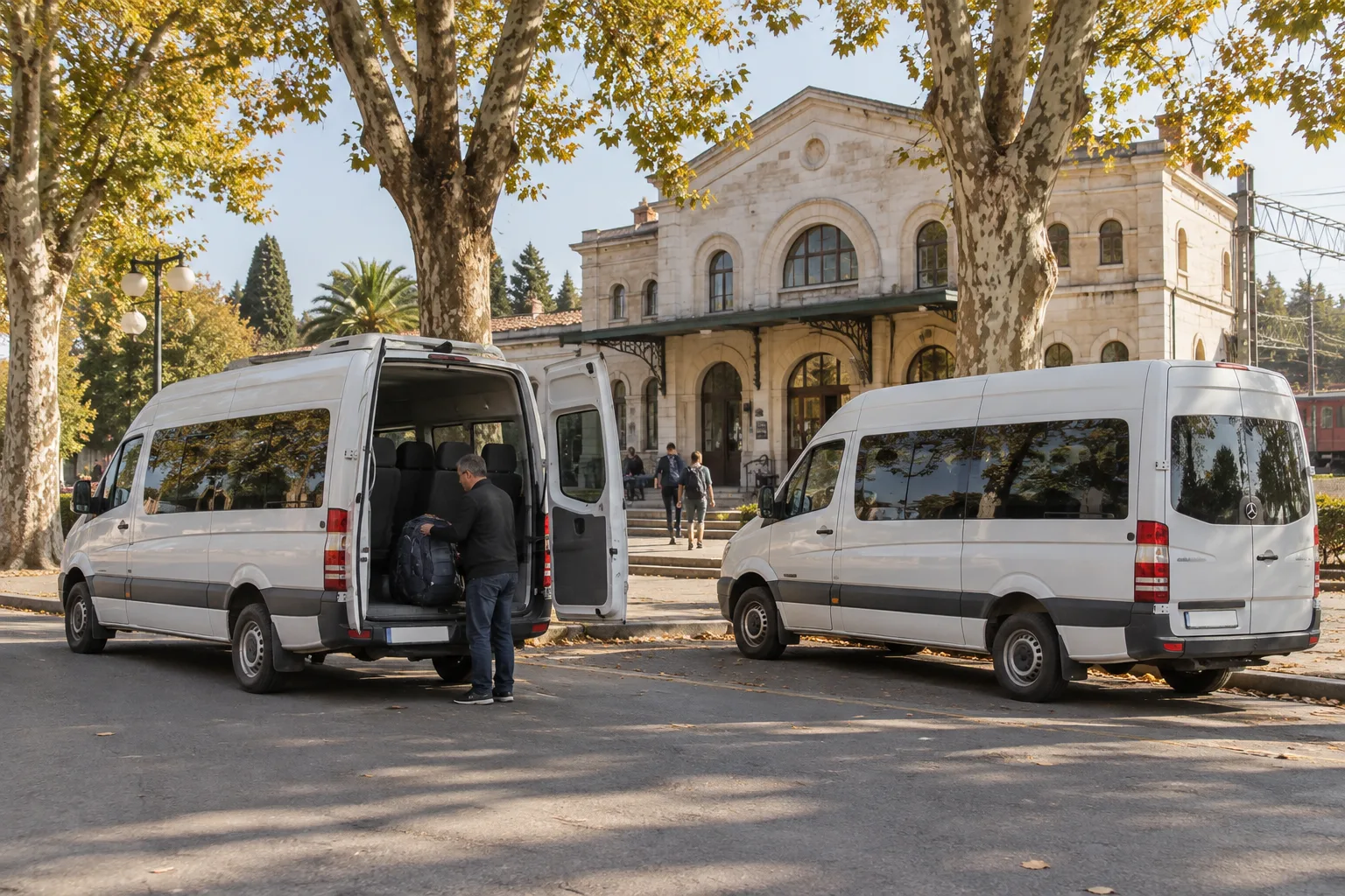 Two white minibus vans parked on a street outside a small provincial railway station in Zugdidi with a driver loading a backpack into the rear door under plane trees