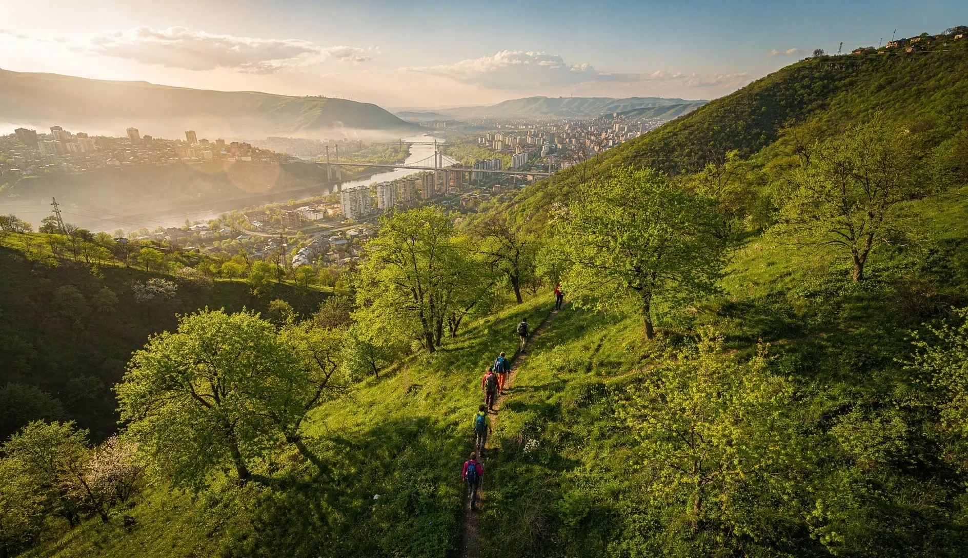Hikers on a forested trail above Tbilisi with the city visible in the valley below during golden afternoon light
