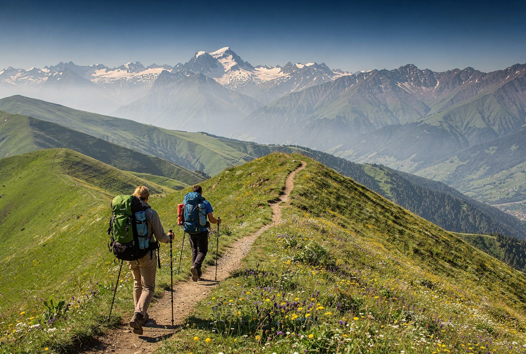 Hikers walking along a grassy ridge trail with panoramic views of green hills and distant Caucasus mountains