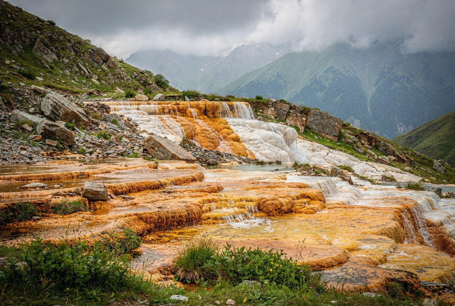 Orange and white travertine mineral deposits along a hillside in Truso Valley with misty Caucasus mountains in the background