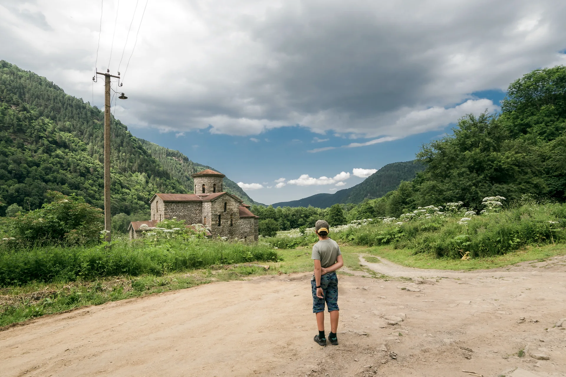 Person standing on a mountain road looking at a medieval stone church in a green Caucasus valley