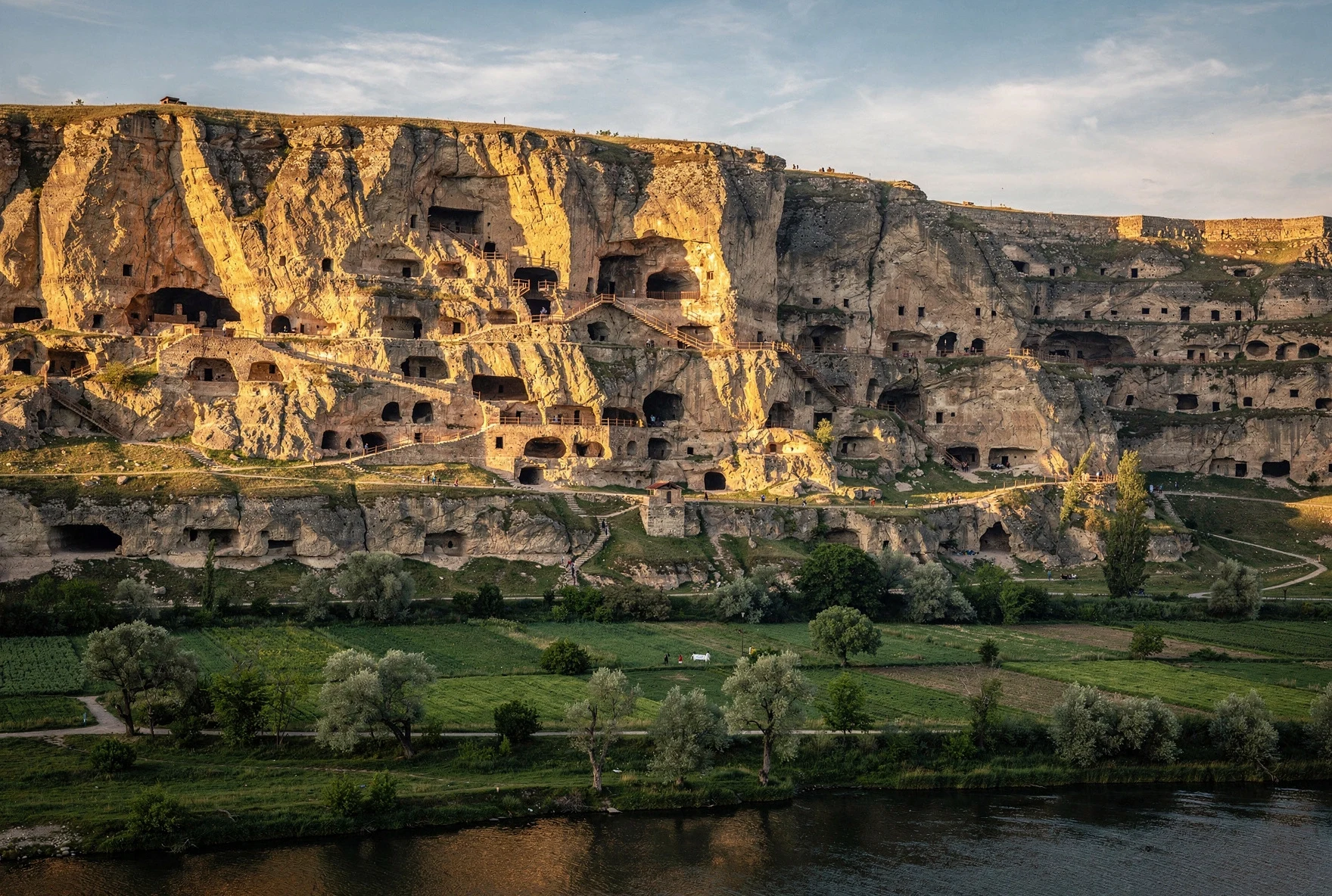 Massive cliff face with hundreds of cave openings carved into rock, green valley below with river