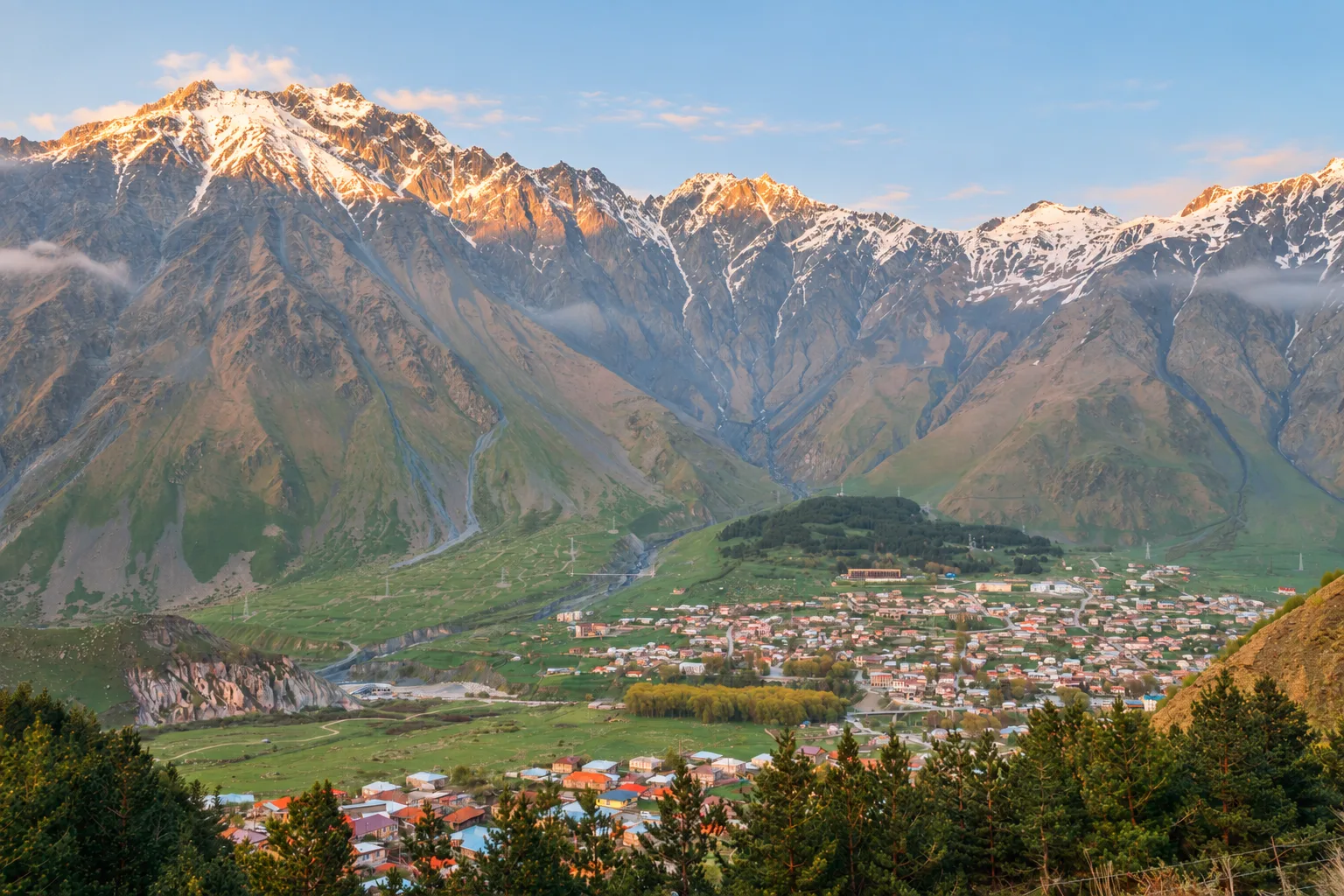 Stepantsminda from the hillside above town on a clear morning, the river meadows and the snow-capped ridge of the Caucasus behind
